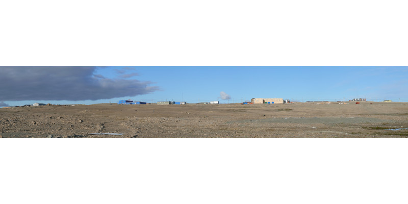 Im Hintergrund stehen einige Hallen und Häuser, vorne ist die karge Landschaft der Arktis zu sehen. 