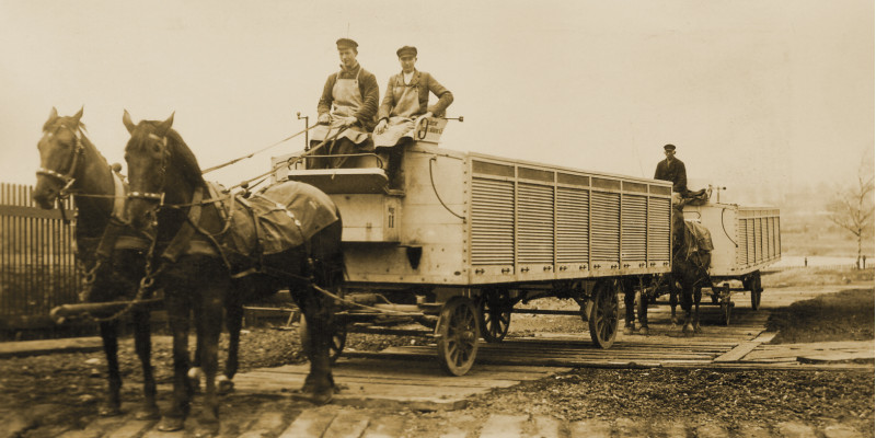 Pfedewagen mit zwei Arbeitern auf dem Kutschbock, die Ladefläche ist mit Metall-Rolläden abgedeckt. 