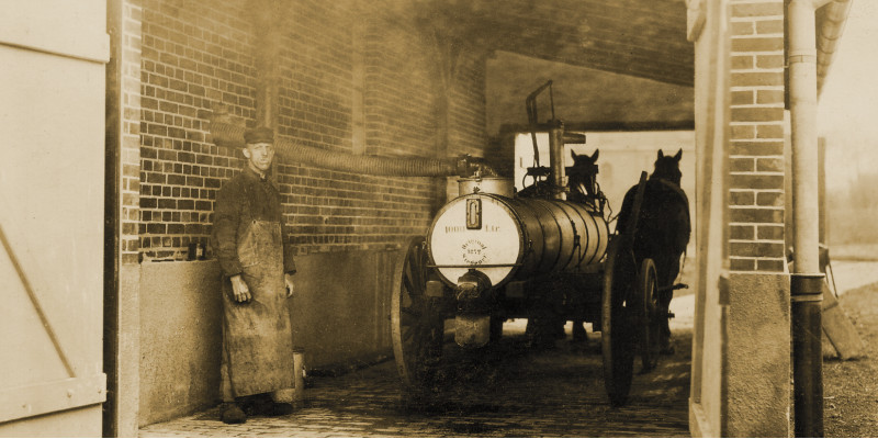 Ein Mann steht mit einem Jauchewagen in einer Einfahrt, der Wagen trägt einen Metalltank und wird von zwei Pferden gezogen. 