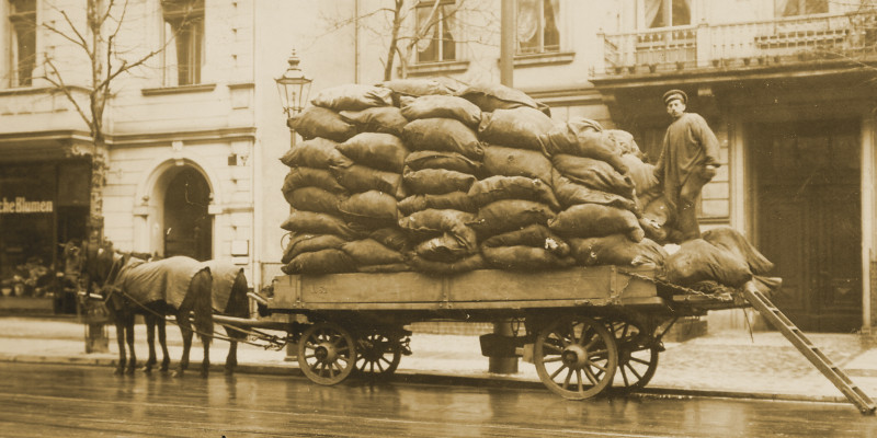 Ein Arbeiter steht auf der Ladefläche eines Pferdewagens, auf der viele Säcke gestapelt sind. Davor steht eine Leiter, über die er auf den Wagen gekommen ist. 