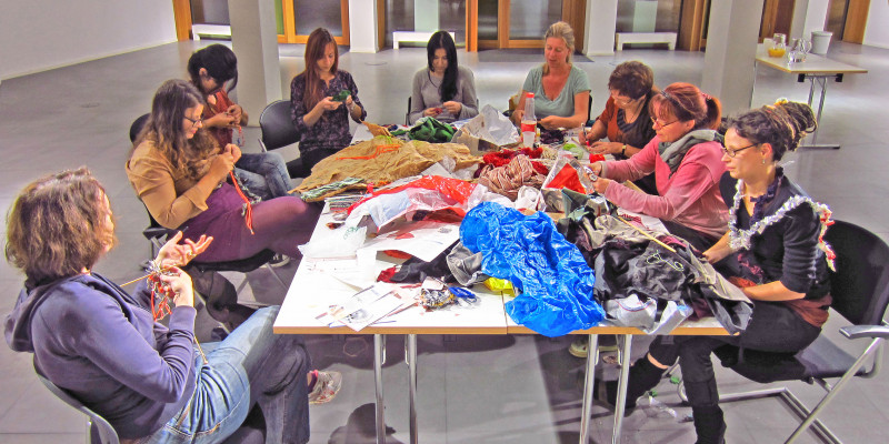 Neun Frauen sitzen an einem Tisch und arbeiten mit Wolle, Stoff und Müll