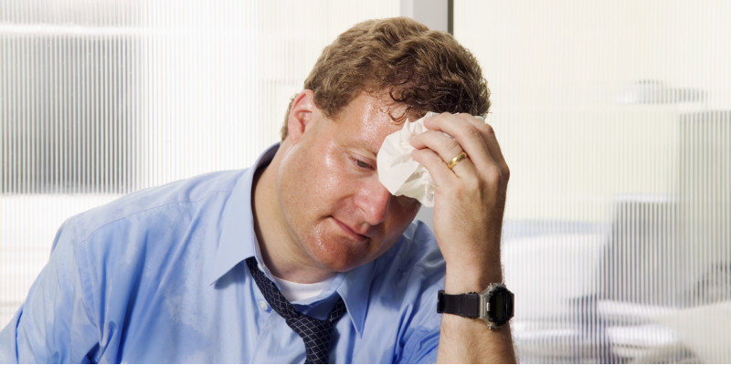 A sweaty man sits in his office in a shirt and tie, wiping his brow.