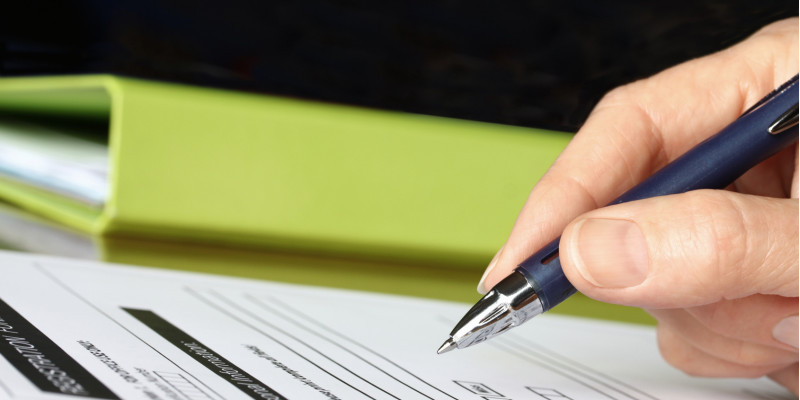 Im Vordergrund Hand hält Stift vor einem Blatt Papier. Im Hintergrund ein grüner Ordner. 