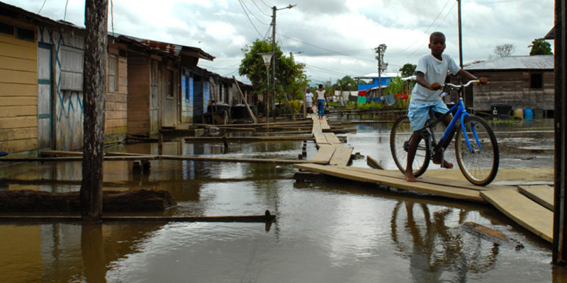 Überschwemmtes Wohngebiet in Entwicklungsland, schwarzes Kind mit Fahrrad auf einem provisorisch angelegten Steg.