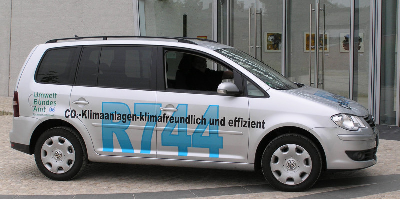 Der silberne klimafreundliche VW Touran, auf dem in großer blauer Aufschrift steht: "R744" und darüber kleiner und schwarz:"CO₂-Klimaanlagen - klimafreundlich und effizient". 