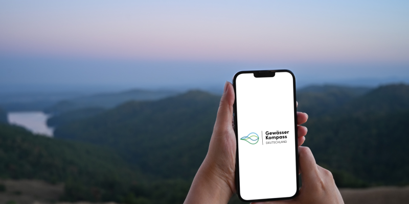 Ein Smartphone wird an einem Fluss benutzt. Auf dem Bildschirm ist zu lesen: "Gewässerkompass Deutschland".