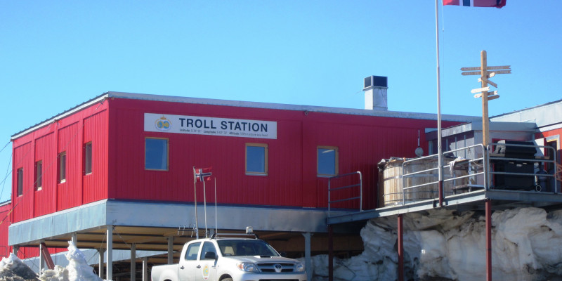 Ein Pick-Up mit Messgeräten auf der Ladefläche steht vor der norwegischen Antarktisstation Troll. Die Station steht auf Stelzen und ist rot. Der Name steht an der Vorderseite auf einem großen Schild. Vor dem Eingang weht die norwegische Flagge.