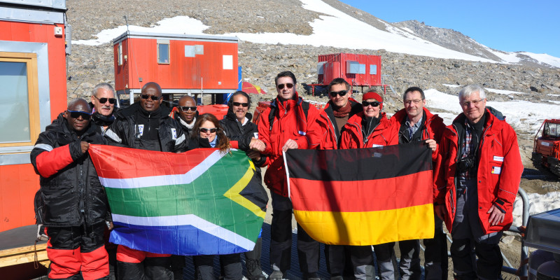 Vor einer Forschungsstation steht eine Gruppe von Männern und Frauen. Links die Gruppe aus Südafrika, recht die deutsche Gruppe. Beide halten ihre jeweilige Landesflagge in den Händen.