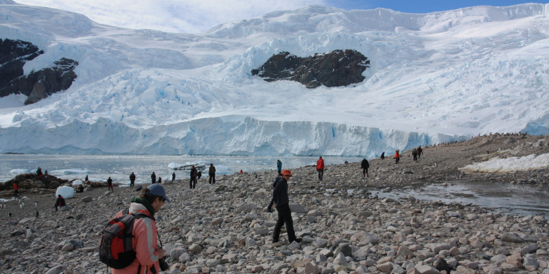 Im Vordergrund laufen Touristen in Funktionskleidung auf steinigem Strand. Im Hintergrund sieht man große Eismassen.