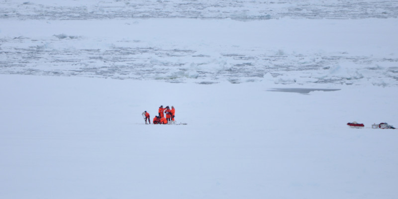 Eine Gruppe von Forschern in orangenen Kälteschutzanzügen steht auf einer großen Eisfläche. Rechts von ihnen, am Bildrand steht ihre Ausrüstung.