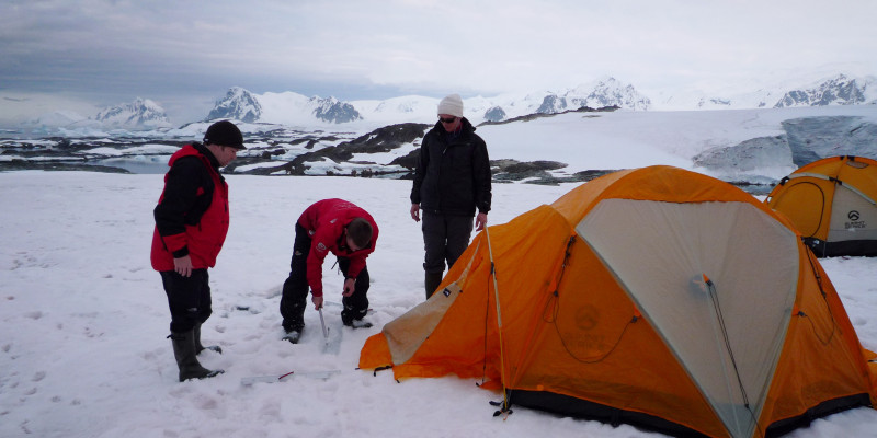 Vor einem in der Antarktis aufgebautem Zelt stehen drei Personen. Sie versuchen die Heringe in den eisigen Boden zu treiben. 