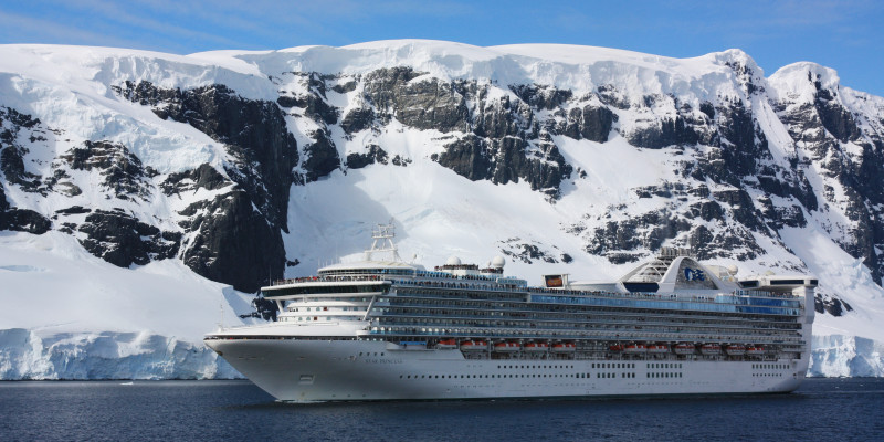 Vor einer schnee- und eisbedeckten Felsenwand fährt ein Kreuzfahrtschiff. Das Wasser ist ruhig, der Himmel ist klar. 