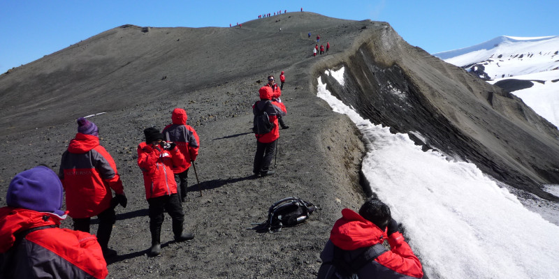 Touristen in roten Funktionsjacken erklimmen einen Hügelgrat. Der Hügel ist nur zur Hälfte schneebedeckt. Einige Leute machen Photos.