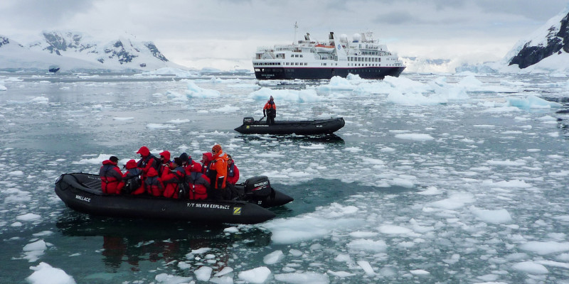 Das antarktische Meer ist mit vielen kleinen Eisschollen und Eisstücke bedeckt. Im Wasser fahren zwei Zodiacs. Im Hintergrund wartet das Kreuzfahrtschiff. 