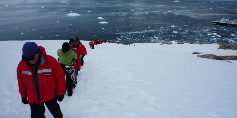 Auf der linken Seite des Bildes kommen Menschen eine schneebedeckte Höhe hochgelaufen. Sie laufen in einer Reihe hintereinander.  Auf dem Meer sieht man das Schiff von dem aus sie gestartet sind. 