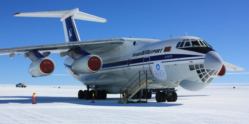 Ein weißes Transportflugzeug mit blauen Streifen an der Seite steht auf der Start- und Landebahn in der Antarktis. Eine Treppe steht vor der Tür. Im Hintergrund fährt ein Pick-up.