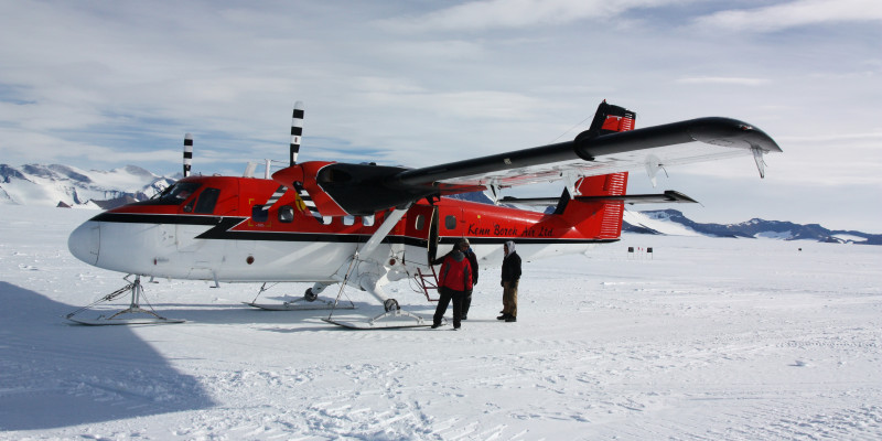 Eine rot-weiße Propellermaschine mit drei Kufen anstelle eines Fahrwerks mit Rädern steht auf einer weiten Schneefläche. Im Hintergrund sind Berge. Vor der Maschine stehen drei Personen. 