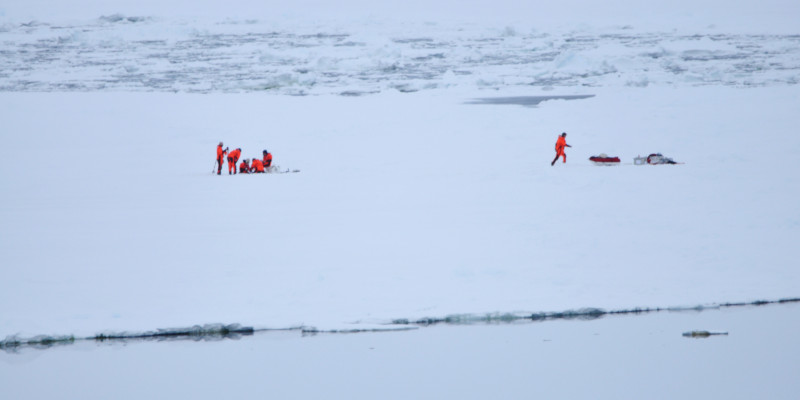 Auf einer weiten Eisebene steht eine Gruppe von Forschern und roten Kälteschutzanzügen. Sie entnehmen an einer Stelle eine Eisprobe. Weiter rechts läuft eine weitere Person zur Ausrüstung. 