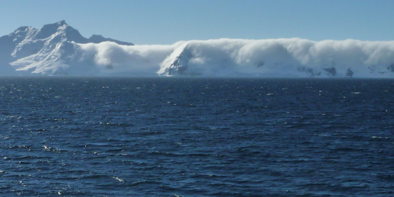 im Vordergrund des Bildes sieht man ruhiges antarktisches Meer. Im Hintergrund des Bildes schließen Berge an, die fast vollständig von Wolken eingehüllt sind. 
