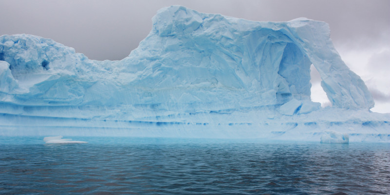 An appealing light blue iceberg floats in the nearly black water. The sky is gray and drab.