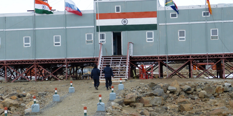 Im Bild ist die indische Forschungsstation auf deren Dach der Name der Station, "Maitri" steht. Davor stehen die Flaggen 4 anderer Länder. " Personen laufen auf die Station zu. 