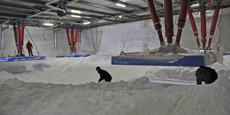Drei Menschen arbeiten unter der Station Neumayer-III, um die Hydraulikstützen mit Schnee zu unterfüttern. Zwei Stelzen sind angehoben. 