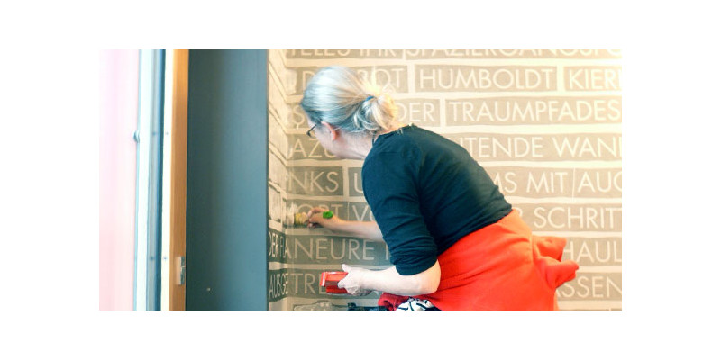 Christiane Keppler streicht mittels weißer Farbe durch die Wortschablonen ihre Texte an die Wand des UBAs Treppenhaus
