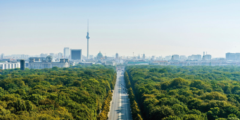 The picture shows an aerial photograph of the ‘Straße des 17. Juni’, a street commemorating the building of the Berlin Wall (the anniversary of German unification takes place annually on 17th June). To the right and left of this street, you can see the canopy of trees in the Große Tiergarten park. The city’s silhouette is visible in the picture’s horizon, including the Reichstags building, tv tower, cathedral and the Brandenburg Gate.