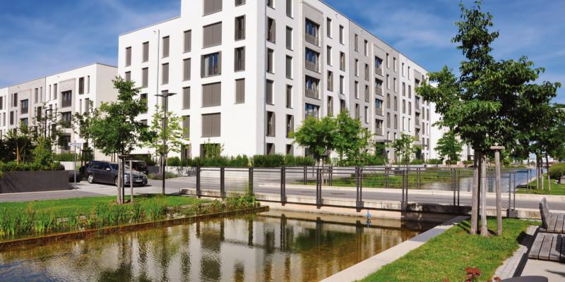 The picture shows an artificial watercourse which runs past a block of flats in a newly developed housing estate. There is a road crossing the watercourse. The watercourse is edged with a narrow strip of vegetation consisting of grass, young trees and flower beds. Benches offer seating. 