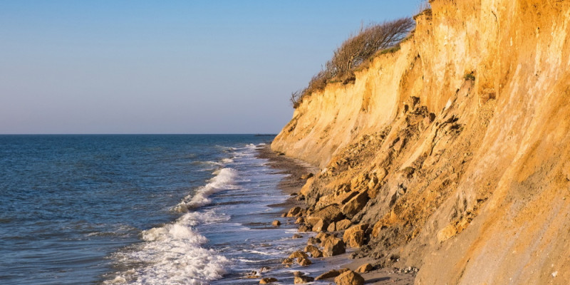 The picture shows a light-brown cliffscape near Ahrenshoop on the Baltic Sea coast. At the foot of the cliff, you can see the sea and the spray of a wave. In one coastal section, you can see loose material and rock fragments which have been shifted from the cliff top to the bottom of the cliff.