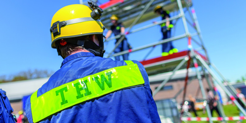 Das Bild zeigt einen Mitarbeiter des Technischen Hilfswerks in Uniform und mit Helm von hinten. Im Hintergrund ist ein Gerüst zu erkennen, auf dem weitere Mitarbeitende des THW stehen.