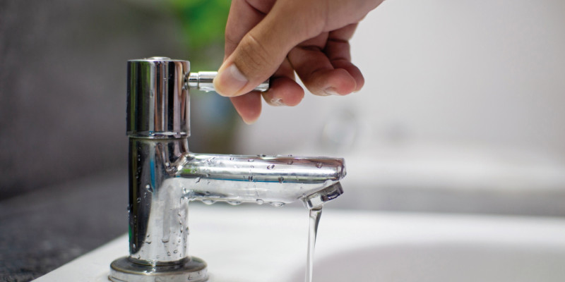 The picture shows a hand on the handle of a tap, with a weak water jet pouring into a wash-hand basin.