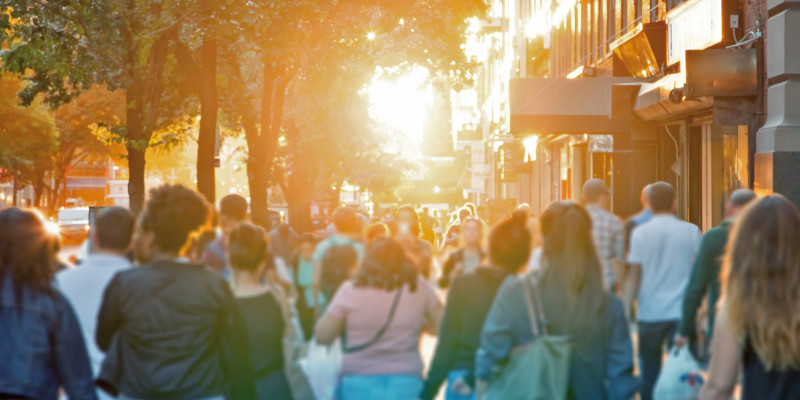 The picture shows an urban pavement alongside a densely built-up road in summer. There are several individuals visible from behind walking on the pavement, some of them carrying shopping bags. The scenery is enveloped in a warm, yellowish light from the low sun. 