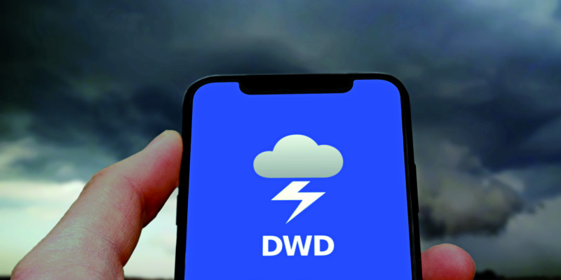 Das Bild zeigt eine Hand, die ein Smartphone hält. Der Bildschirm des Smartphones ist blau und zeigt das Logo der WarnWetter-App des DWD. Im Hintergrund des BIldes sind die schwarzen Wolken eines Gewitterhimmels zu erkennen.