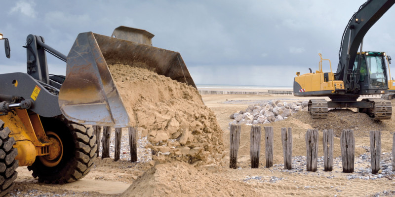 Das Bild zeigt im Vordergrund die gefüllte Schaufel eines Radladers, aus der Sand auf einen Sandstrand entleert wird. Im Hintergrund sind hölzerne Pfähle und eine Steinhalde auf einem weiten Sandstrand zu sehen. Der Himmel ist von dunkelgrauen Wolken verhangen.