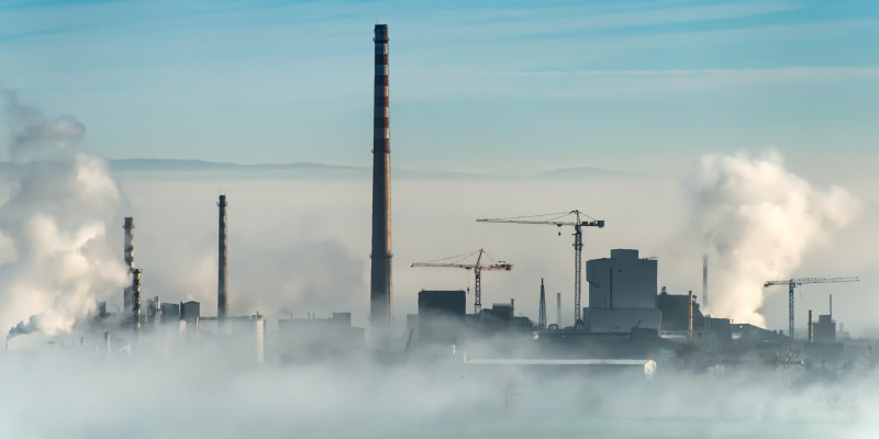 Industrieanlage mit mehreren Rauchtürmen und zwei Baukränen auf dem Gebiet; im Vordergrund steigt Nebel aus dem Kühlwasserflussauf