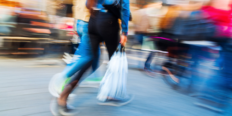 hektischer, verschwommener Hintergrund, der sich anscheinend dreht; im Mittelpunkt stehen zwei Frauen, die durch die Fußgängerzone schlendern und Einkaufstüten in ihren Händen tragen