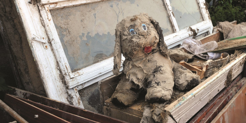 The picture shows piled up heavily soiled boards and windows. On top of the pile is a cuddly toy dog completely covered in mud.