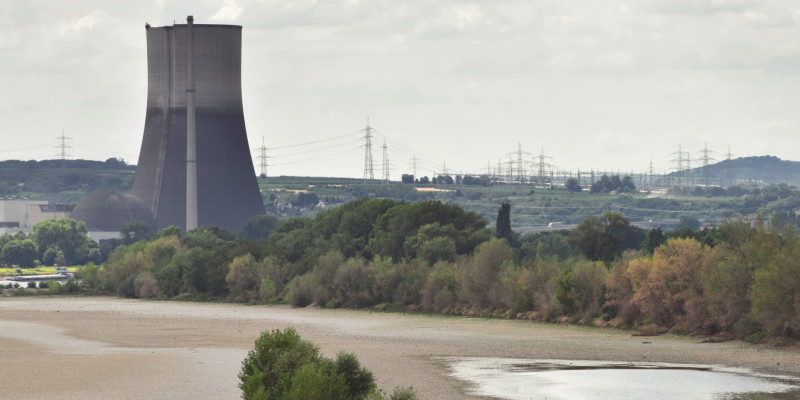 The picture shows a riverbed that has almost dried up and is lined with rows of trees. A cooling tower rises in the near background.