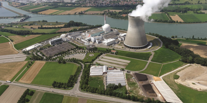 The image shows an aerial view of a nuclear power plant with a cooling tower emitting water vapour at the edge of a river.