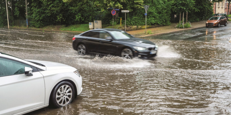 Das Bild zeigt eine völlig überflutete Straßenkreuzung in einer Stadt, in die gerade zwei Autos hineinfahren.