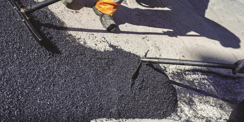 The picture shows the legs of a road construction worker who is repairing a piece of road with fresh asphalt.