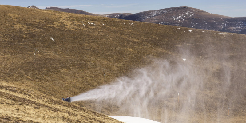 Das Bild zeigt eine in Betrieb befindliche Schneekanone in einem bergigen Pistengelände, auf dem kein Schnee liegt. 