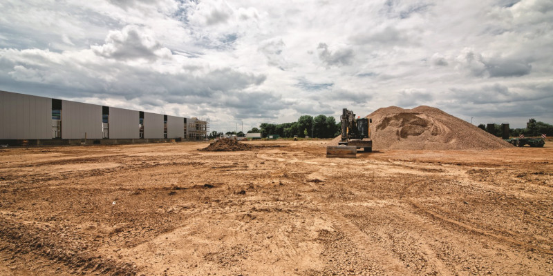 Das Bild zeigt eine große offene Bodenfläche auf einer Baustelle. Im Hintergrund steht rechts neben einem aufgeschütteten Erdhügel ein Bagger auf der Fläche, links sieht man Teile einer Industriehalle. 