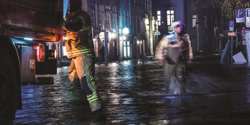 Das Bild zeigt einen mit Wasser überfluteten Straßenzug bei Nacht. Zwei Einsatzkräfte waten durch das Wasser, einer davon befindet sich direkt an einem Einsatzfahrzeug. 
