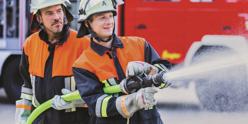 Das Bild zeigt einen jungen, freudestrahlenden Feuerwehrmann, der einen Spritzschlauch betätigt. Hinter ihm steht ein älterer Feuerwehrmann, der ihn offensichtlich anleitet. 