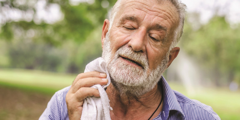 The picture shows an elderly man with closed eyes drying his neck with a sweat towel.