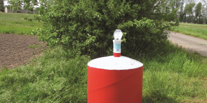 The picture shows a groundwater measuring point in the landscape. A nozzle protrudes from a round red base.