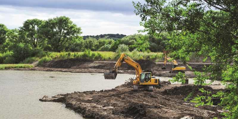 The picture shows a water body on the edge of which a construction measure is being carried out. The edge of the bank is open ground where two dredgers are active.