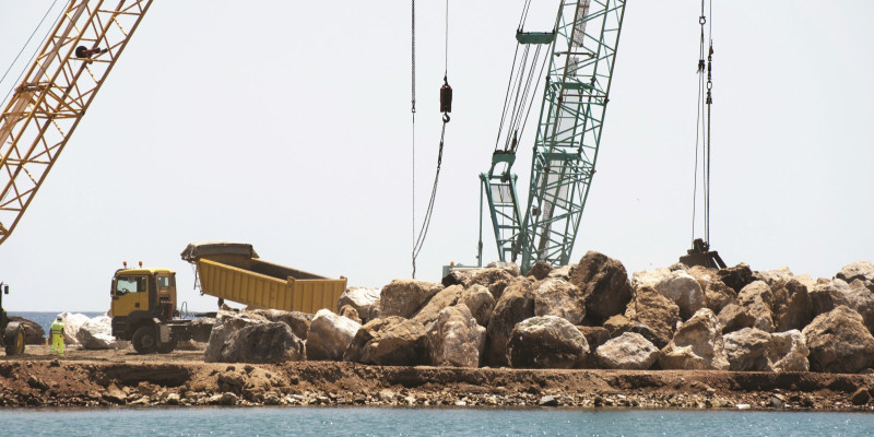 The picture shows a large construction project by the sea. Large blocks of stone are being moved by large cranes and a large truck. 